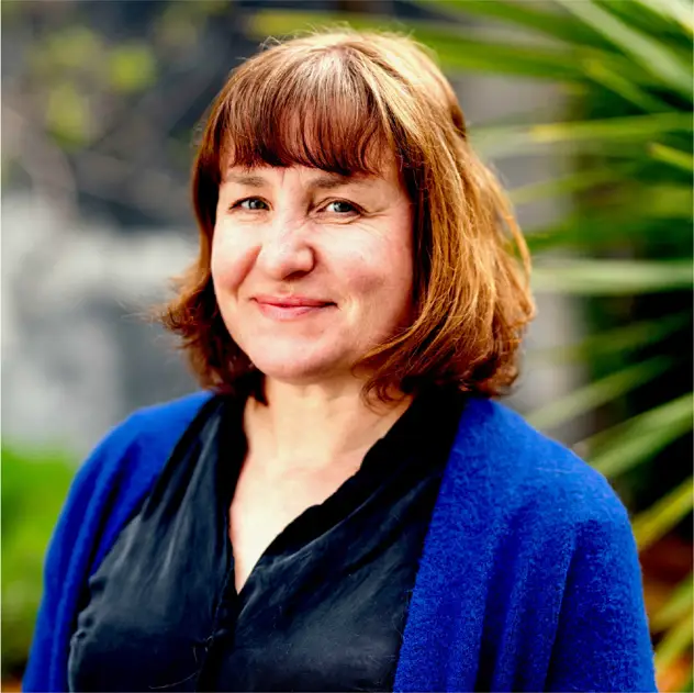 A woman with shoulder-length brown hair and bangs, wearing a blue cardigan over a black top, stands outdoors with greenery in the background.