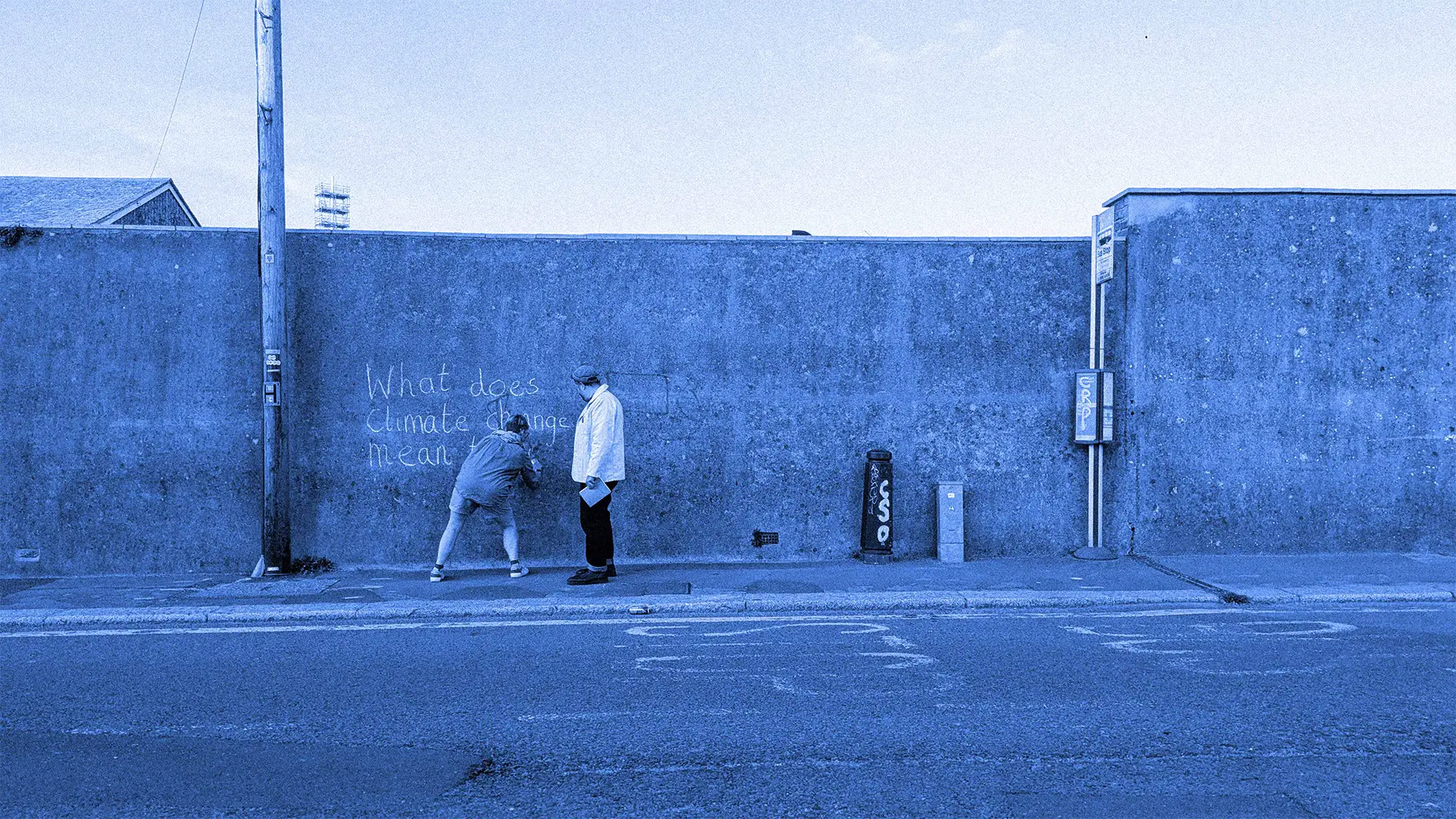 Two people standing next to a concrete wall outdoors, with a clear sky above. They are writing the words "What Does Climate Change Mean" in chalk.
