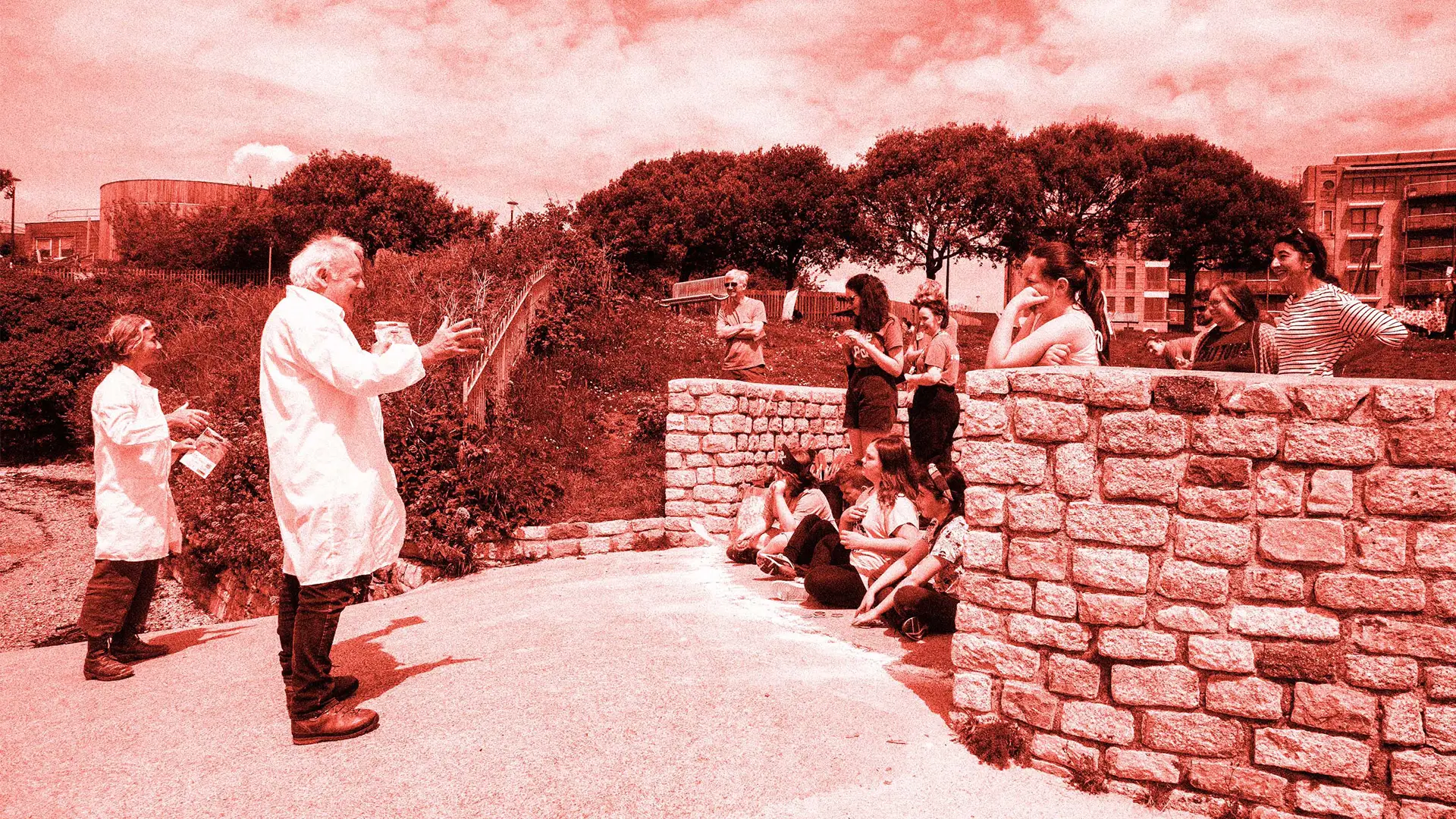 A man and woman in lab coats stand in front of a stone wall, surrounded by a group of people. The scene takes place outdoors under a cloudy sky, with trees and buildings in the background. Everyone is dressed casually and appears to be interacting or enjoying the moment together.